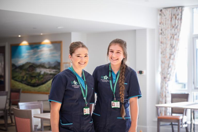 Two young women wearing care uniform smile towards the camera