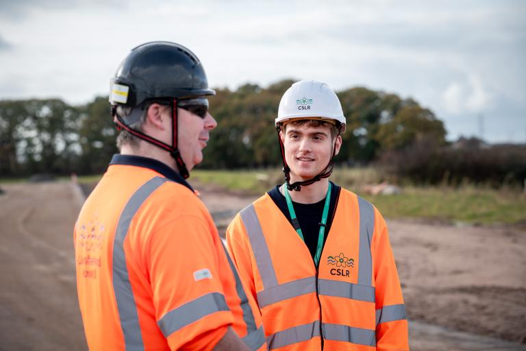 Two men wear orange hi-vis jackets marked CSLR, the younger man looks like he is listening to the older man